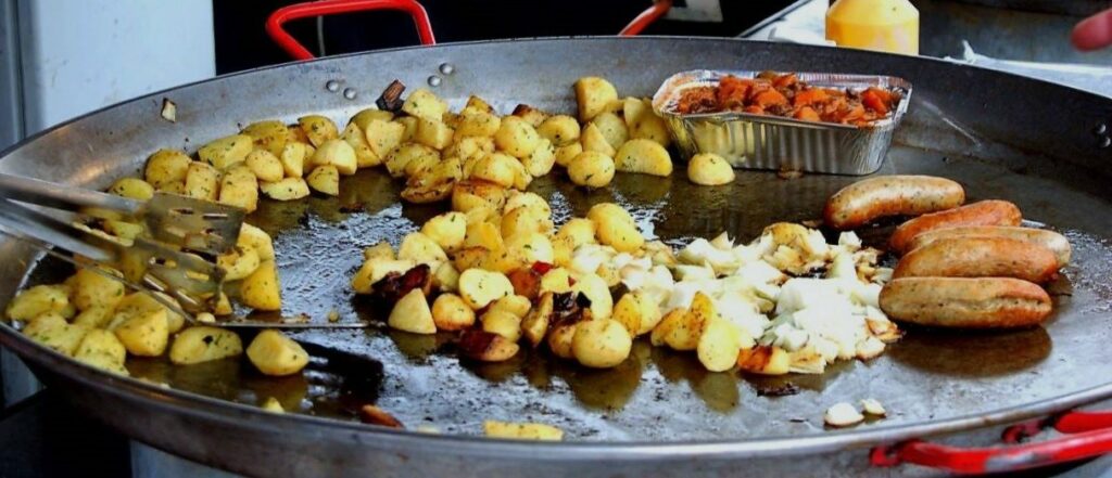 large pan at a food festival stall with sausages and hash brown being cooked on it