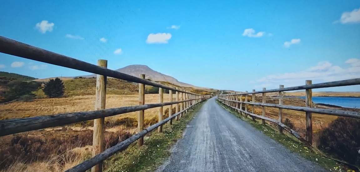 clew bay bike trail on a sunny day, blue sky, fields on either side of the road with wooden fencing.