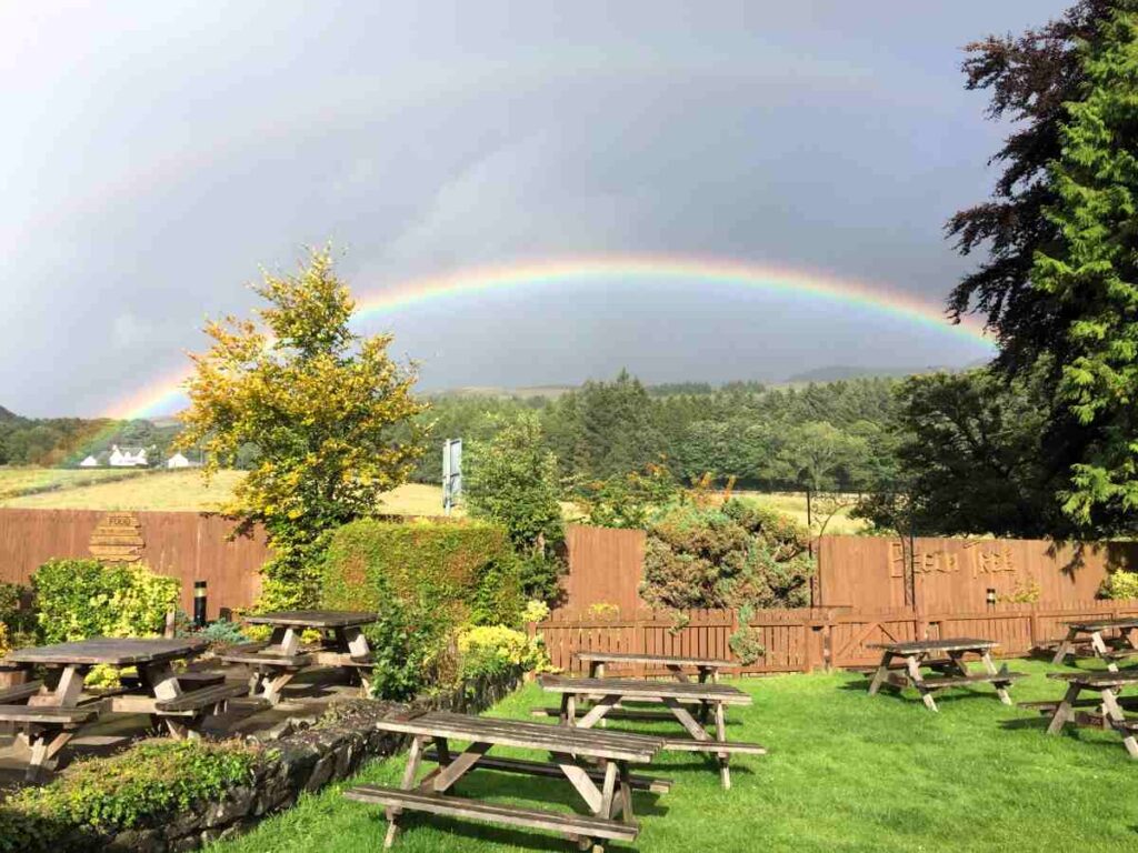 Rainbow stretching between hills in the distance with red roofs underneath and picnic tables in the foreground, at the beech tree in Milngavie to drymen hike