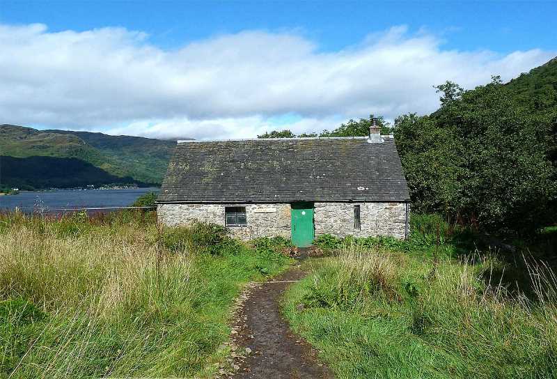 Photo of Doune Bothy - simple stone cottage among long grasses with an earth path towards its green door