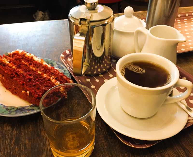 Table set with a pot of tea, tea cup, slice of chocolate cake, glass of whiskey, milk and sugar bowls