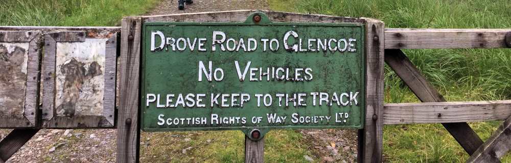 Green metal sign on a wooden gate at the start of a hike through Rannoch Moor, writing says 'Drove Road To Glencoe No Vehicles, Please keep to the track'