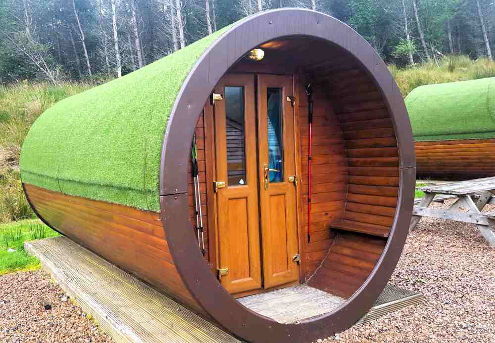 long rounded wooden cabin with green artificial grass covering its top part. A microloge in Glencoe Mountain Resort on West Highland Way